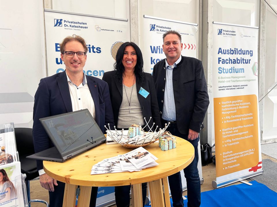 Drei Personen stehen lächelnd hinter einem runden Holztisch auf einer Messe. Auf dem Tisch liegen Flyer, ein Laptop und kleine Giveaways. Hinter ihnen sind Roll-up-Banner der „Privatschulen Dr. Kalscheuer“ mit Informationen zu Ausbildung, Fachabitur und Studium in den Bereichen E-Business Management sowie Hotel- und Tourismusmanagement zu sehen. Die Szene wirkt freundlich und einladend.