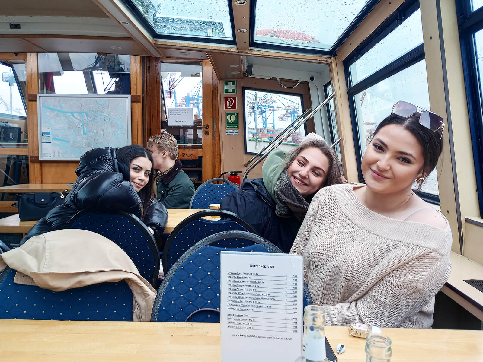 Vier junge Menschen sitzen in einem Innenraum mit großen Fenstern, vermutlich auf einem Ausflugsschiff oder in einem Café. Drei von ihnen schauen lächelnd in die Kamera, während eine Person im Hintergrund zur Seite blickt. Auf dem Tisch im Vordergrund steht eine Getränkekarte, daneben leere Flaschen. Durch die Fenster sind Hafenkräne und regnerisches Wetter zu sehen.
