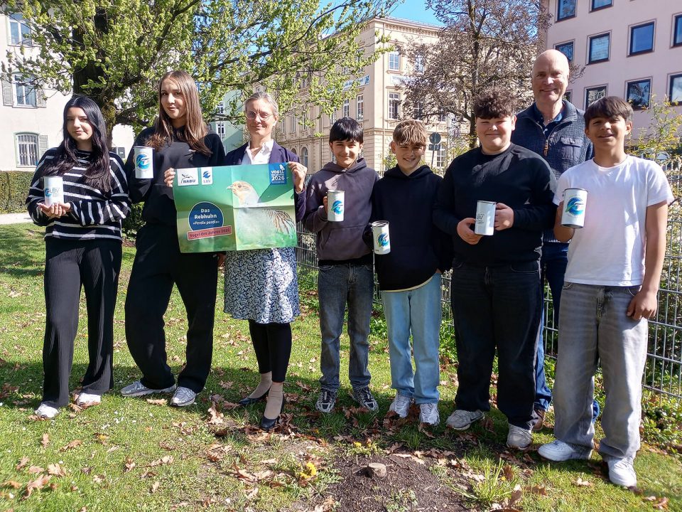 Sieben Personen stehen nebeneinander auf einer Rasenfläche im Freien. Alle halten weiße Tassen mit einem blauen Logo. Eine Person in der Mitte hält zusätzlich ein großes rechteckiges Schild mit einem Foto eines Vogels und einem runden blauen Logo. Im Hintergrund sind mehrstöckige Gebäude, Bäume mit frischem Laub und ein Metallzaun zu sehen. Die Personen tragen überwiegend dunkle Kleidung.