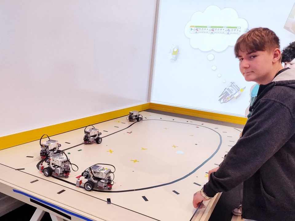 Ein Junge mit braunen Haaren steht an einem Tisch mit erhöhtem gelbem Rand. Auf der Tischoberfläche fahren mehrere kleine, weiß-graue Roboter mit sichtbaren Kabeln entlang einer schwarzen Linie. Im Hintergrund ist eine weiße Wand mit einer Illustration zu sehen, die eine Sprechblase und einen gezeichneten Roboter zeigt.