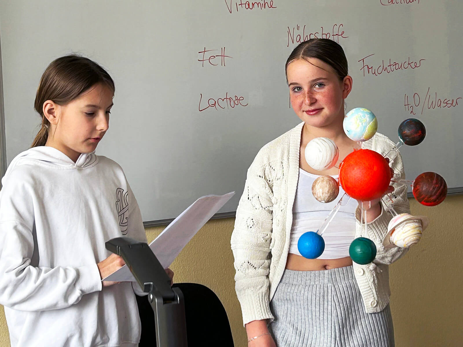 Zwei Kinder stehen vor einer Tafel im Klassenraum. Links liest ein Kind aus einem Blatt und hält ein Tablet vor sich. Rechts hält ein anderes Kind ein selbstgebasteltes Modell des Sonnensystems mit bunten Kugeln, das um eine große rote „Sonne“ angeordnet ist. Auf der Tafel im Hintergrund stehen handschriftliche Begriffe wie „Vitamine“, „Fett“, „Lactose“, „Nährstoffe“, „Fruchtzucker“ und „H₂O/Wasser“.