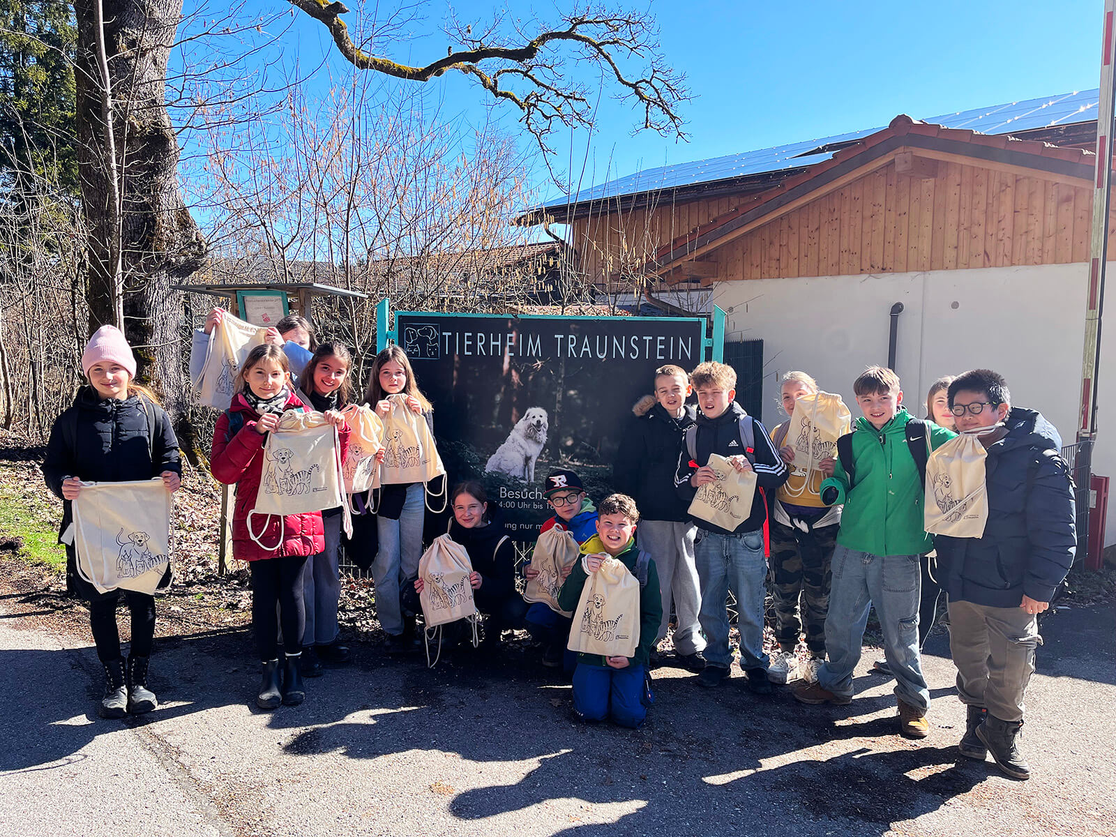 Eine Gruppe von Kindern steht im Halbkreis vor einem großen Schild mit der Aufschrift 'TIERHEIM TRAUNSTEIN' und dem Bild eines Hundes. Die Kinder halten Stoffbeutel mit sichtbaren Aufdrucken in den Händen. Im Hintergrund sind ein Gebäude mit Holzfassade und ein kahler Baum zu sehen. Die Szene spielt bei sonnigem Wetter im Freien.