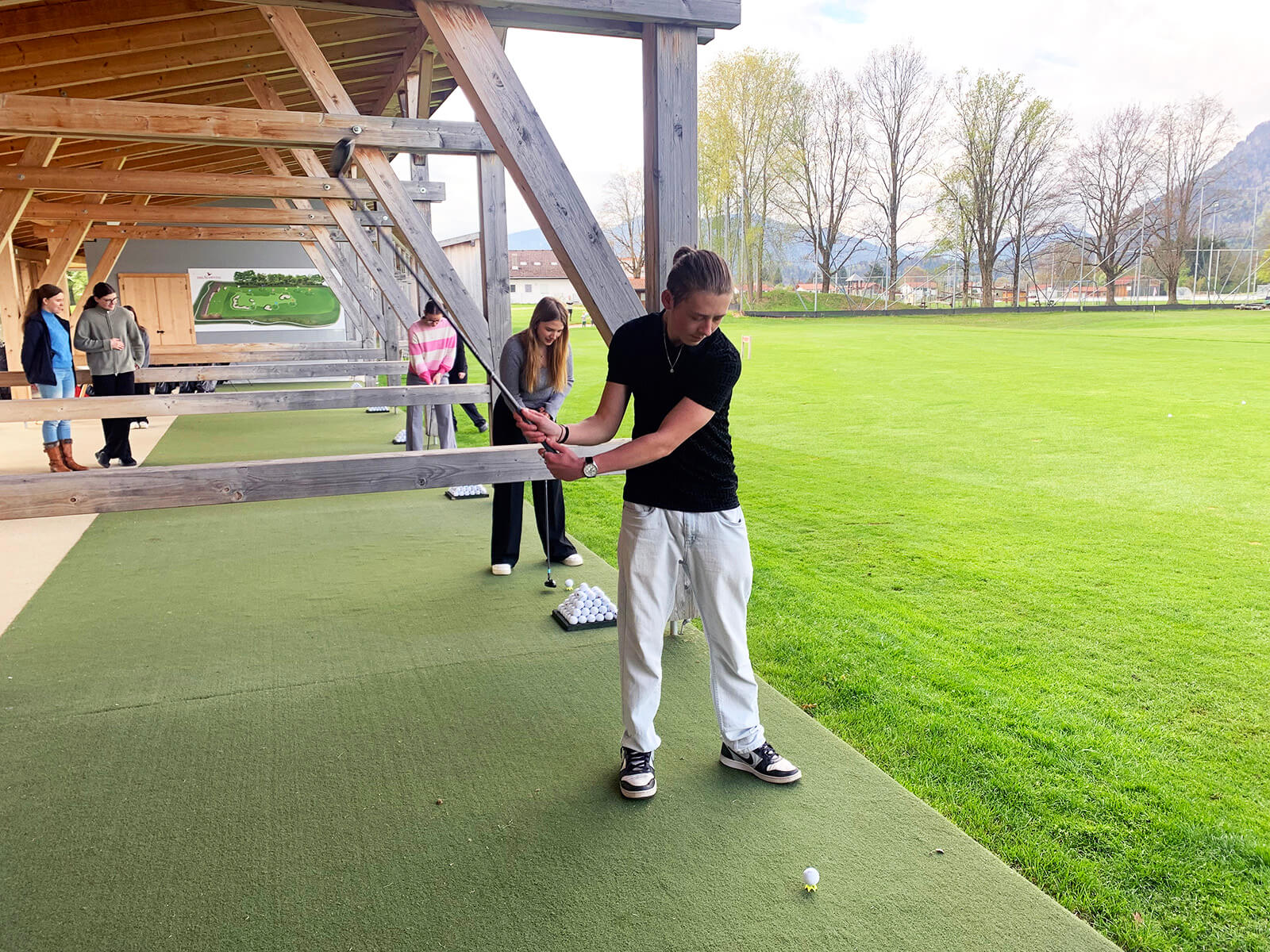 Eine Person steht auf einer überdachten Abschlagmatte und bereitet sich mit einem Golfschläger auf einen Abschlag vor. Im Hintergrund stehen weitere Personen, die ebenfalls Golfschläger halten oder beobachten. Die Szenerie befindet sich an einer gepflegten, weitläufigen Rasenfläche mit Bäumen und Bergen im Hintergrund. Die Personen tragen sportliche Freizeitkleidung.
