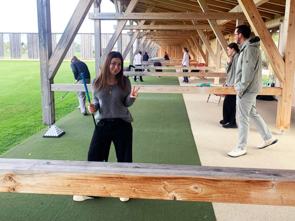 Überdachte Golf-Driving-Range mit Holzkonstruktion. Im Vordergrund steht eine Person mit langen Haaren, grauem Pullover und dunkler Hose auf grünem Kunstrasen, hält einen Golfschläger in der rechten Hand und zeigt mit der linken Hand ein Victory-Zeichen. Im Hintergrund üben weitere Personen an den Abschlagplätzen, einige tragen helle Jacken. Rechts verläuft ein schmaler, heller Weg parallel zum Rasen.