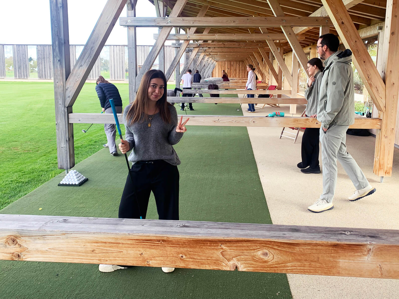 Überdachte Golf-Driving-Range mit Holzkonstruktion. Im Vordergrund steht eine Person mit langen Haaren, grauem Pullover und dunkler Hose auf grünem Kunstrasen, hält einen Golfschläger in der rechten Hand und zeigt mit der linken Hand ein Victory-Zeichen. Im Hintergrund üben weitere Personen an den Abschlagplätzen, einige tragen helle Jacken. Rechts verläuft ein schmaler, heller Weg parallel zum Rasen.