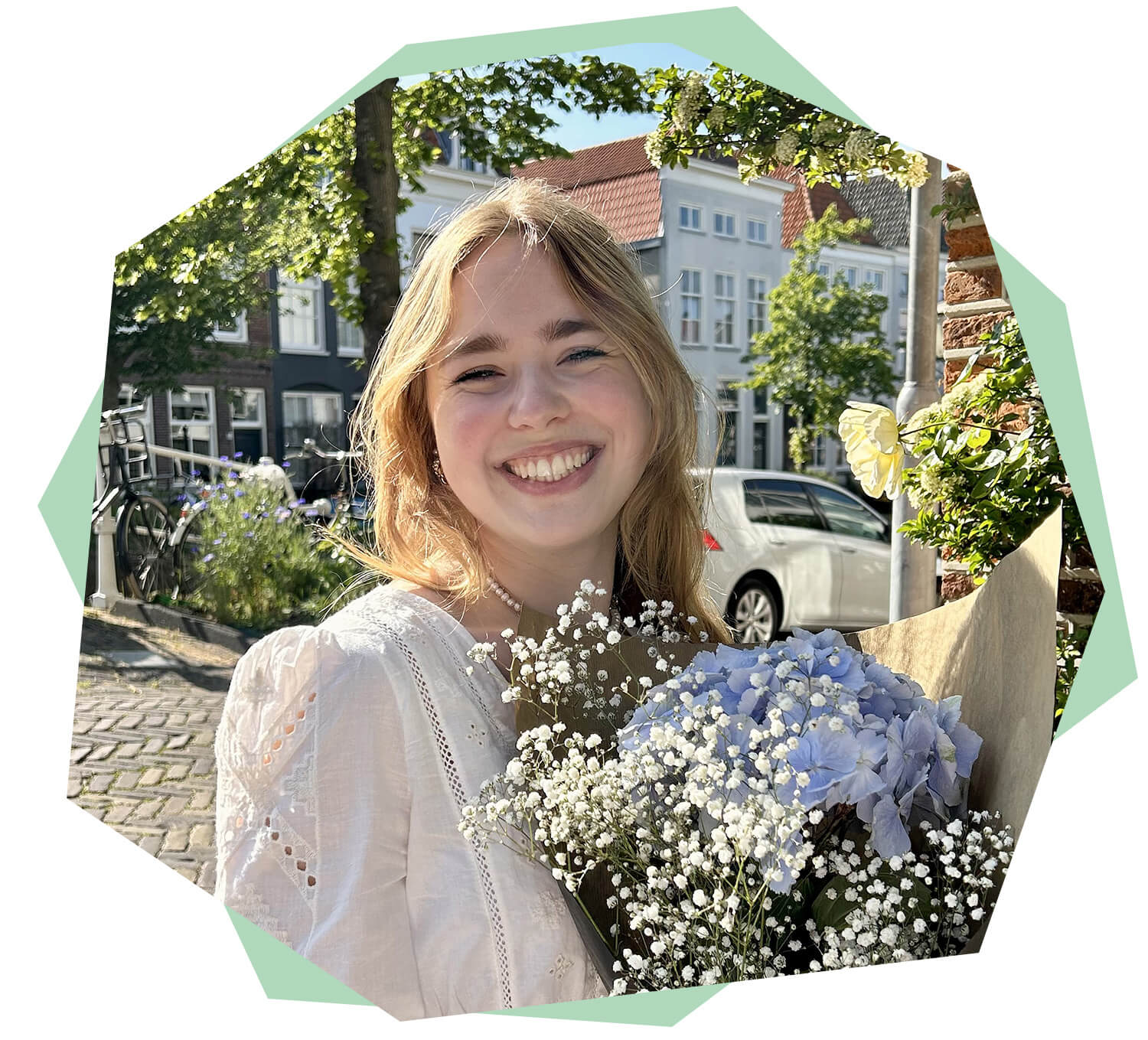 Porträt einer lächelnden jungen Frau im Freien. Sie trägt eine helle Bluse und hält einen Blumenstrauß mit weißen und hellblauen Blüten in den Armen. Im Hintergrund sind sonnige Bäume, Fahrräder und Häuser entlang einer ruhigen Straße zu sehen.