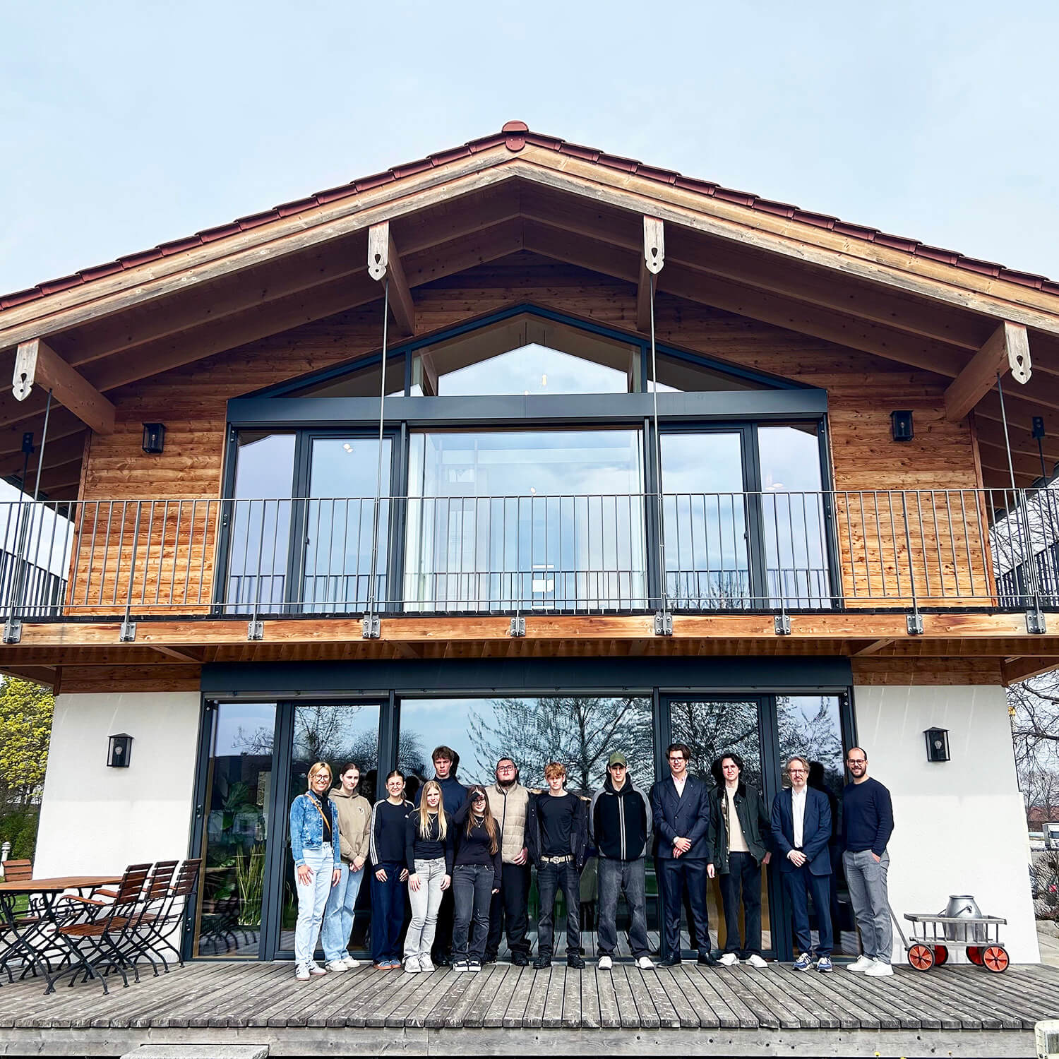 Vor einem modernen Holzhaus mit großem Balkon und bodentiefen Fenstern steht eine Gruppe von Menschen in lockerer Reihe. Das Gebäude hat eine Fassade aus Holz und Glas, ein Satteldach mit roten Ziegeln und sichtbare Holzbalken. Links und rechts neben dem Eingang stehen Gartenmöbel. Der Himmel ist hell und wolkenlos.