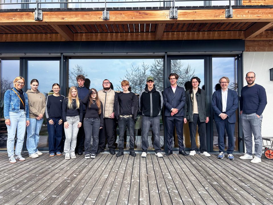 Eine Gruppe von Menschen steht auf einer Holzterrasse vor einer modernen Glasfassade. Die Personen sind in einer Reihe aufgestellt, einige mit verschränkten Armen, andere mit den Händen in den Taschen oder locker an den Seiten. Die Kleidung reicht von leger bis formell, darunter Jacken, Pullover, Hemden und Anzüge. Im Hintergrund spiegeln sich Bäume in den großen Fenstern. Über der Gruppe ist ein Balkon mit Metallgeländer und Holzbalken zu sehen. Rechts am Bildrand steht ein Kinderwagen.