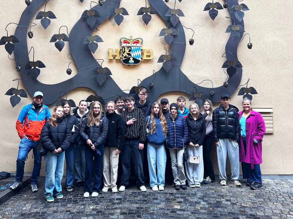 Eine Gruppe Jugendlicher und Erwachsener steht auf nassem Kopfsteinpflaster vor einer beigen Wand. An der Wand ist ein großes, dekoratives Metallornament mit Blättern und Hopfendolden befestigt. In der Mitte des Ornaments befindet sich ein Wappen mit bayerischen Rauten und den goldenen Buchstaben 'HB'. Die Personen tragen überwiegend Freizeitkleidung und Jacken, einige haben Kapuzen oder Mützen auf.