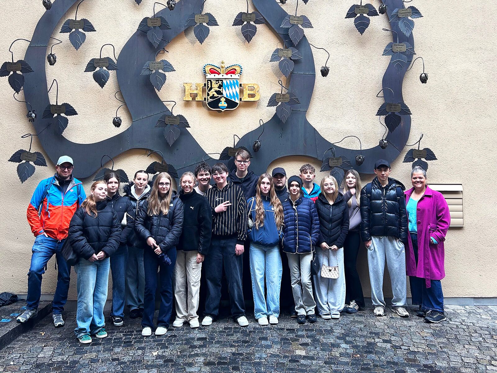 Eine Gruppe Jugendlicher und Erwachsener steht auf nassem Kopfsteinpflaster vor einer beigen Wand. An der Wand ist ein großes, dekoratives Metallornament mit Blättern und Hopfendolden befestigt. In der Mitte des Ornaments befindet sich ein Wappen mit bayerischen Rauten und den goldenen Buchstaben 'HB'. Die Personen tragen überwiegend Freizeitkleidung und Jacken, einige haben Kapuzen oder Mützen auf.