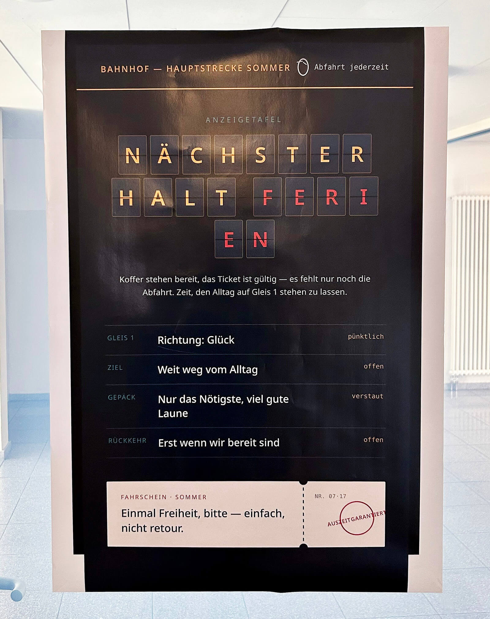 Im Mittelpunkt steht eine große, schwarze Anzeigetafel mit farbigen Buchstaben, die das Wort 'NÄCHSTER HALT FERIEN' hervorheben. Darüber befindet sich eine weiße Leiste mit der Aufschrift 'BAHNHOF – HAUPTSTRECKE SOMMER Abfahrt jederzeit'. Unter der Hauptzeile sind verschiedene Reiseinformationen wie Ziel, Richtung, Proviant, Rückkehr und ein Fahrtschein abgebildet, die humorvoll auf das Thema Urlaub und Erholung anspielen. Die Tafel steht in einem hellen, modernen Raum mit glänzendem Boden und einem weißen Heizkörper im Hintergrund.