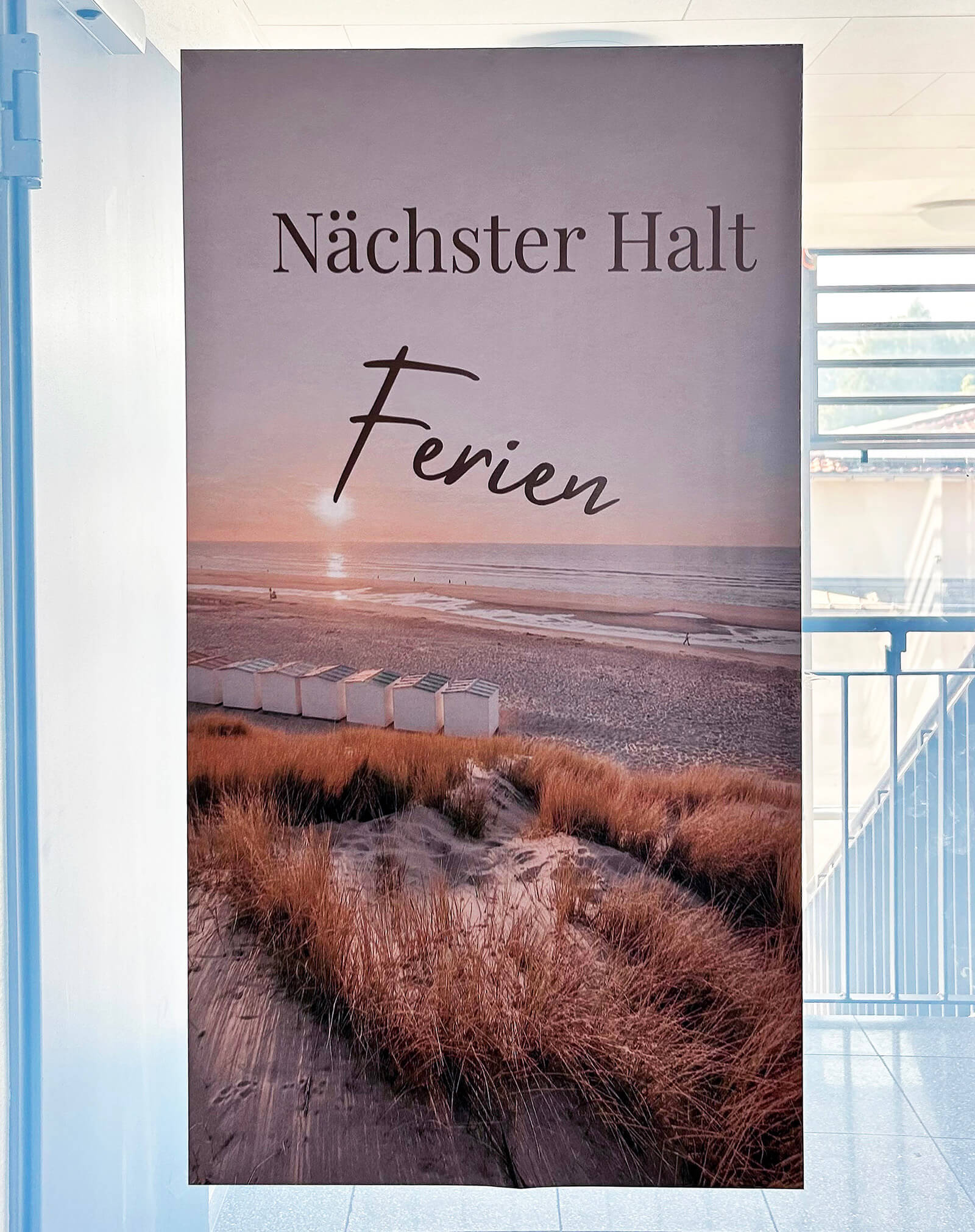 Ein hochformatiges Stoffbanner hängt in einem Flur vor einem Fenster. Das Banner zeigt eine Fotografie von Dünen mit trockenem Gras, einem Holzsteg und einem Strand mit weißen Strandkabinen. Im Hintergrund ist das Meer zu sehen, darüber ein Sonnenuntergang mit warmen Farbtönen. Im oberen Bereich des Banners steht in großer, klarer Schrift: 'Nächster Halt Ferien'.