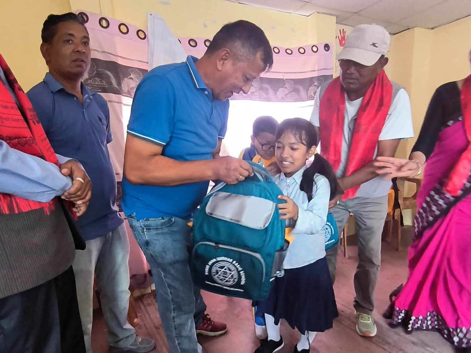 Ein Mädchen in Schuluniform hält einen türkisfarbenen Rucksack mit einem weißen Emblem, während ein Mann im blauen T-Shirt ihr den Rucksack überreicht. Mehrere Erwachsene stehen um das Mädchen herum, einige tragen rote Schals. Im Hintergrund sind weitere Kinder und ein hellgelber Raum mit Vorhängen zu sehen. Die Stimmung wirkt feierlich und unterstützend.