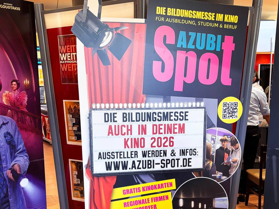 Großes Plakat mit dem Schriftzug 'AZUBI Spot' und einer Leuchttafel mit dem Text 'DIE BILDUNGSMESSE AUCH IN DEINEM KINO 2026 AUSSTELLER WERDEN & INFOS: WWW.AZUBI-SPOT.DE' vor einem Messestand.