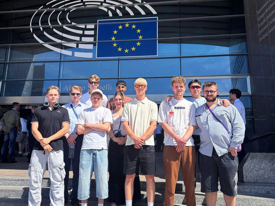 Gruppe junger Menschen steht vor dem Eingang des Europäischen Parlaments mit EU-Flagge und Glasfassade im Hintergrund.