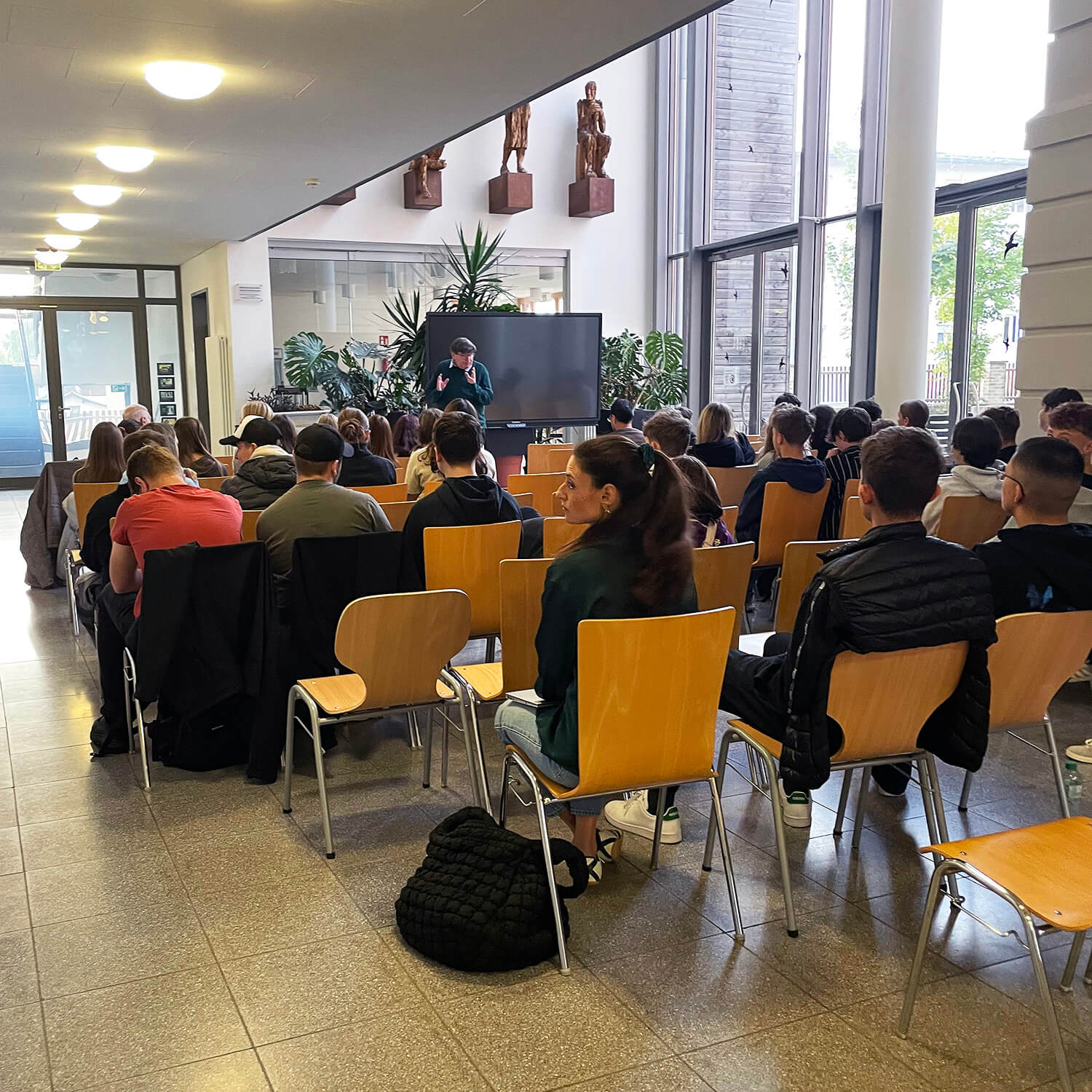 In einem hellen, modernen Raum mit großen Fenstern sitzen zahlreiche Menschen auf hellen Holzstühlen in mehreren Reihen. Im Vordergrund sind Rücken und Profile der Zuhörenden zu sehen, einige blicken zur Seite oder nach vorne. Im Hintergrund steht eine Person vor einem großen Bildschirm und spricht zu den Anwesenden. Pflanzen und Skulpturen sind im hinteren Bereich des Raumes platziert. Das Licht ist gleichmäßig und freundlich.