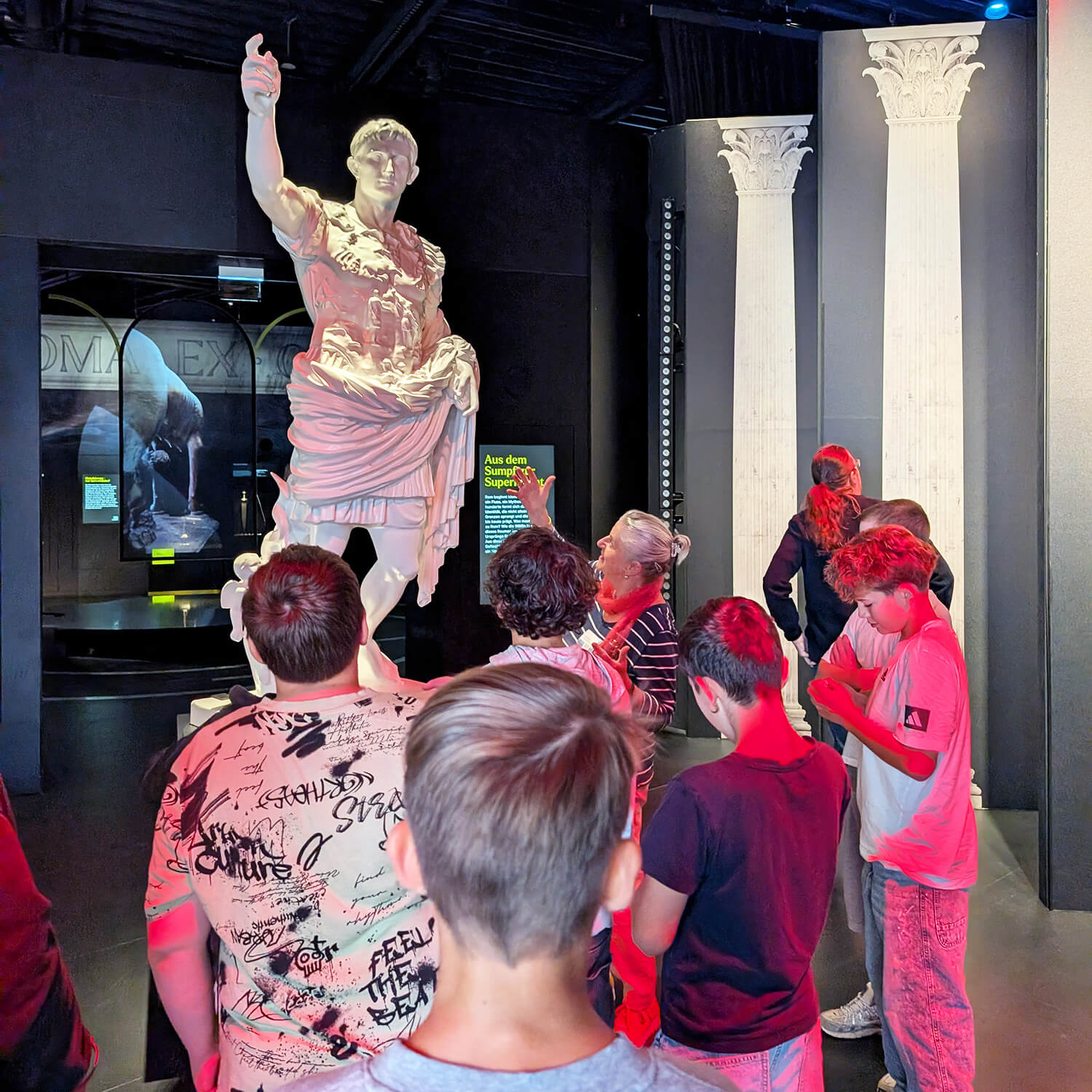 Im Vordergrund stehen mehrere Jugendliche mit dem Rücken zur Kamera, die eine große Statue eines römischen Herrschers betrachten. Die Statue zeigt eine Person in Rüstung mit erhobenem rechten Arm. Im Hintergrund sind zwei weiße Säulen und eine dunkle Ausstellungswand mit beleuchteten Informationstafeln zu sehen. Die Szene ist von farbigem Licht, insbesondere Rot, beleuchtet.