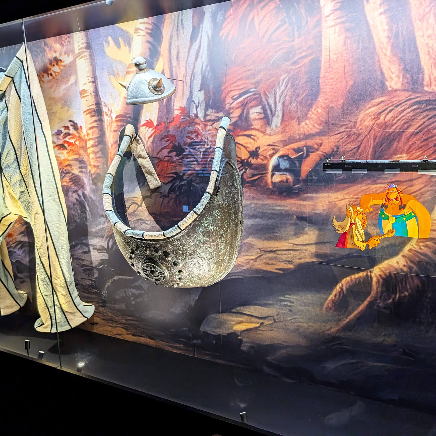 Im Vordergrund sind mehrere Ausstellungsstücke in einer Vitrine zu sehen, darunter ein halbrundes, metallisch wirkendes Objekt mit Verzierungen und ein weiteres, längliches Objekt mit Streifenmuster. Im Hintergrund befindet sich eine farbige Wandgestaltung mit einer Waldszene. Rechts ist eine bunte Zeichnung von zwei Comicfiguren aufgebracht, die an den Stil von Asterix und Obelix erinnert. Die Szene ist hell ausgeleuchtet und die Objekte sind durch Glasscheiben geschützt.