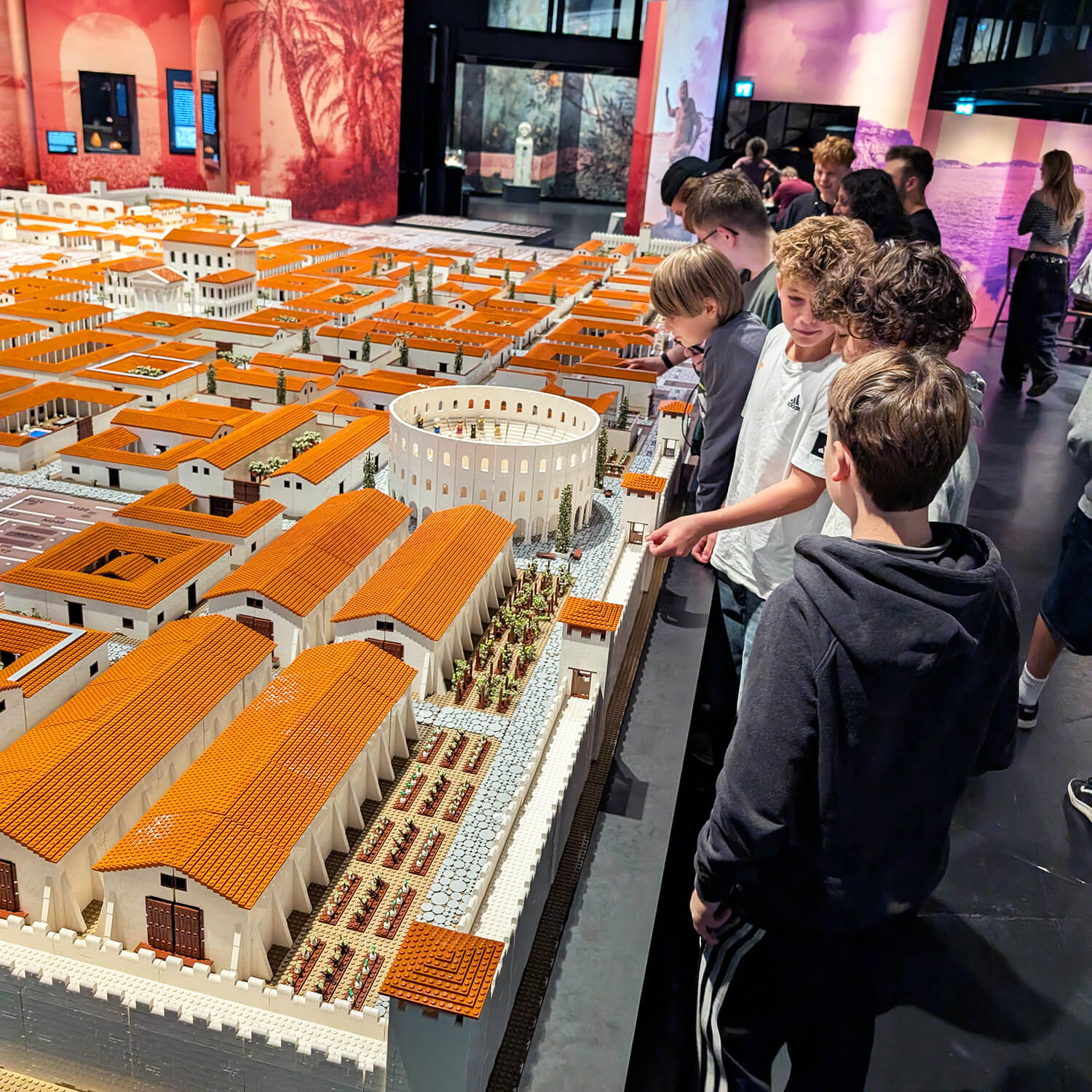Kinder und Jugendliche stehen an einem großen Modell einer antiken römischen Stadt mit orangefarbenen Dächern und weißen Mauern. Im Zentrum des Modells befindet sich ein rundes Amphitheater. Die Szene spielt in einem Ausstellungsraum mit farbigen Wandbildern und moderner Beleuchtung. Einige der Kinder zeigen auf Details des Modells, andere betrachten es aufmerksam.