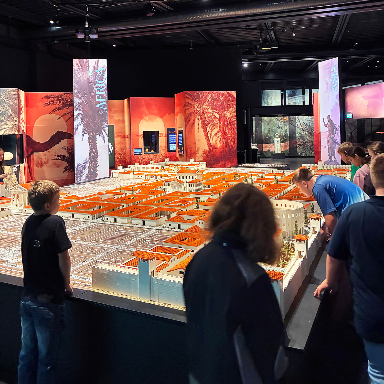 Im Vordergrund betrachten mehrere Personen ein großes Modell einer antiken Stadt mit weißen Mauern und roten Dächern. Das Modell ist detailreich gestaltet und nimmt einen Großteil des Raumes ein. Im Hintergrund sind Ausstellungstafeln mit Palmenmotiven und warmen Farbtönen zu sehen. Die Beleuchtung ist gedämpft, wodurch die Szene einen musealen Charakter erhält.