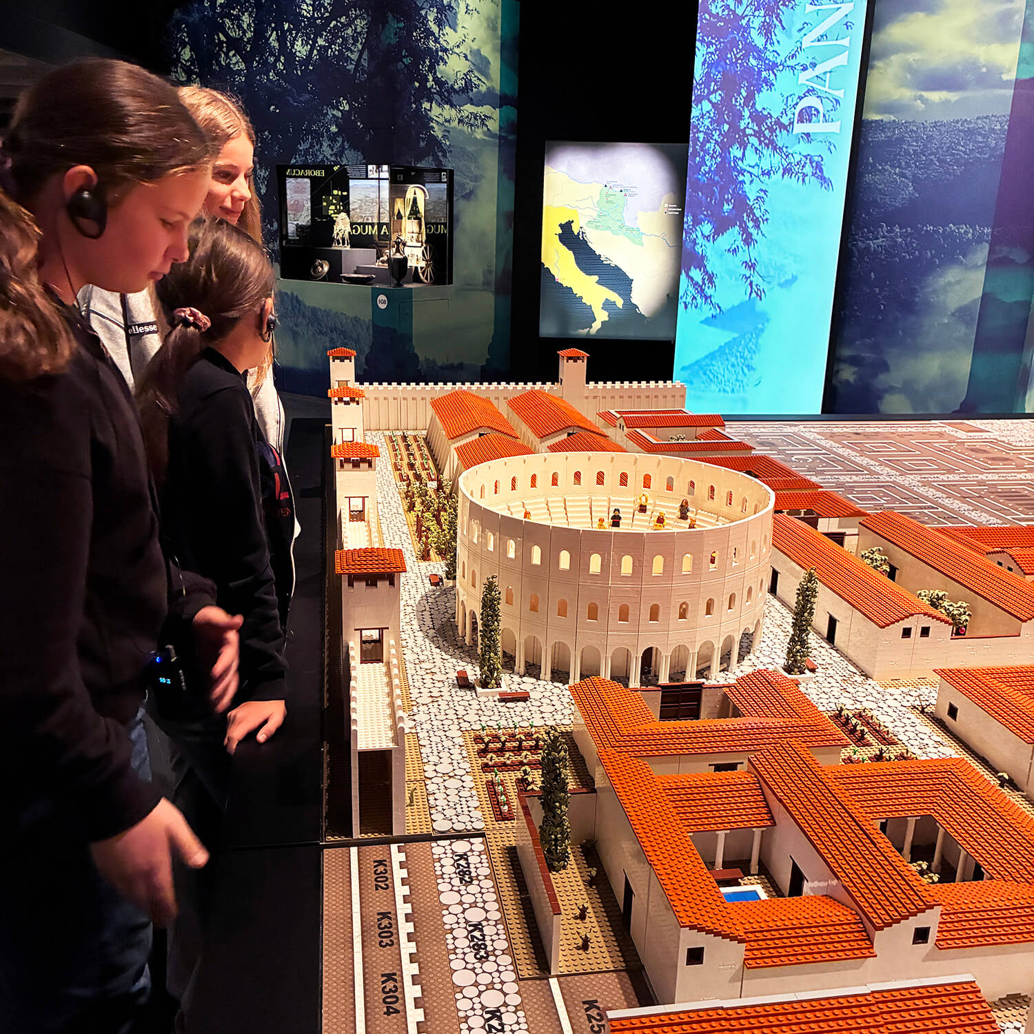 Drei Kinder stehen an einem großen Modell einer römischen Stadt mit einem zentralen Amphitheater. Das Modell zeigt zahlreiche Gebäude mit roten Ziegeldächern, gepflasterte Straßen und kleine Bäume. Die Kinder tragen Kopfhörer und betrachten das detailreiche Stadtmodell aus nächster Nähe. Im Hintergrund sind beleuchtete Ausstellungstafeln mit Karten und Texten zu sehen.