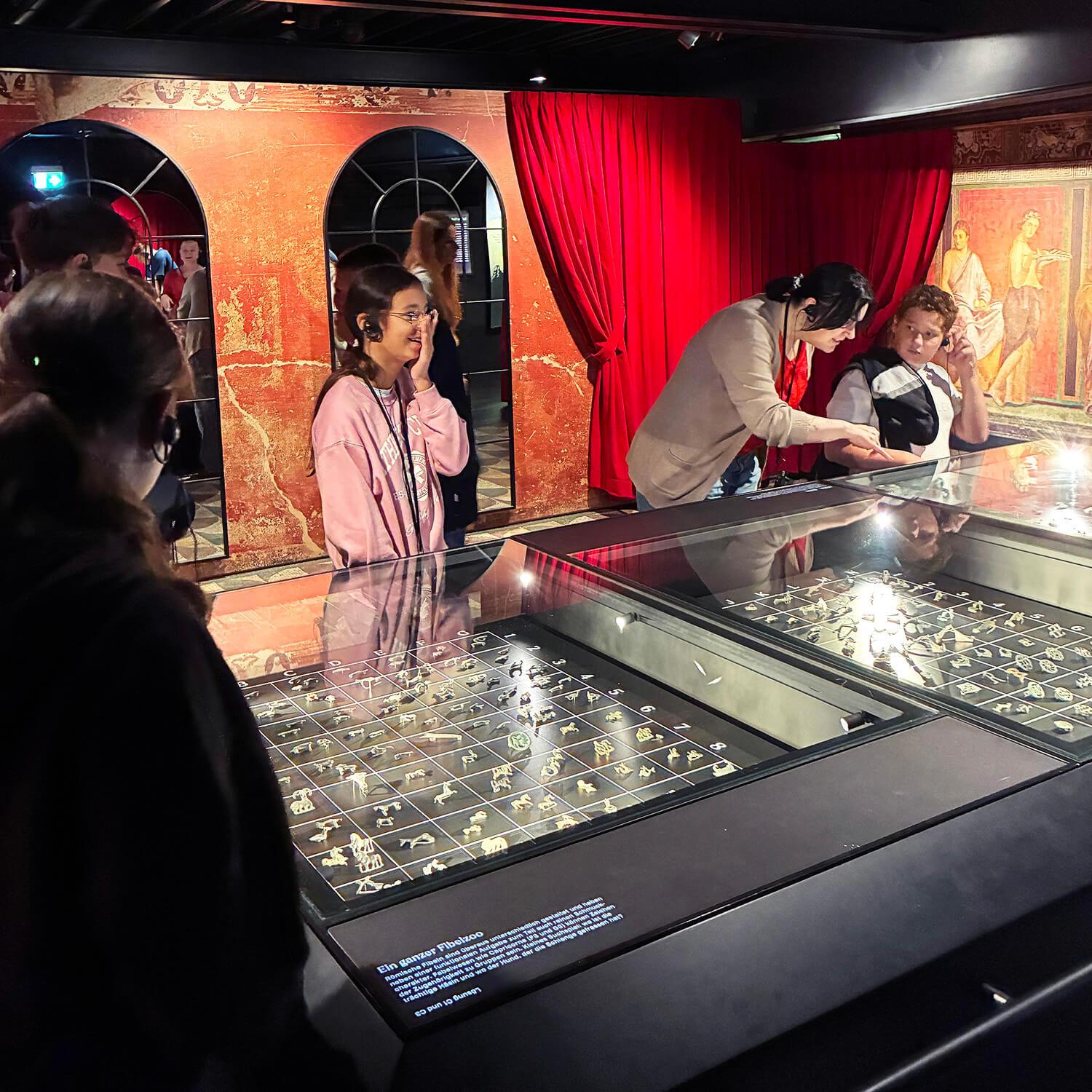 Mehrere Personen stehen in einem abgedunkelten Ausstellungsraum vor beleuchteten Vitrinen, in denen zahlreiche kleine Fundstücke oder Schmuckstücke präsentiert werden. Im Hintergrund sind rote Vorhänge und Wandmalereien mit römischen Motiven zu sehen. Einige Personen tragen Kopfhörer und betrachten die Exponate aufmerksam.