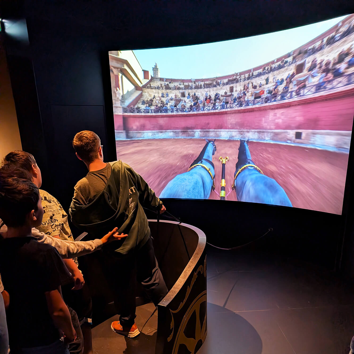 Mehrere Personen stehen an einem interaktiven Ausstellungsobjekt, das wie ein antiker Streitwagen gestaltet ist. Sie blicken auf eine große, gebogene Leinwand, auf der eine virtuelle Szene aus der Perspektive eines Wagenlenkers in einer römischen Arena gezeigt wird. Die Leinwand zeigt die Vorderbeine zweier Pferde und die Tribünen mit Zuschauern. Die Szene ist farbig und vermittelt den Eindruck einer Simulation oder eines Videospiels. Die Umgebung ist dunkel gehalten, sodass der Fokus auf der Projektion liegt.