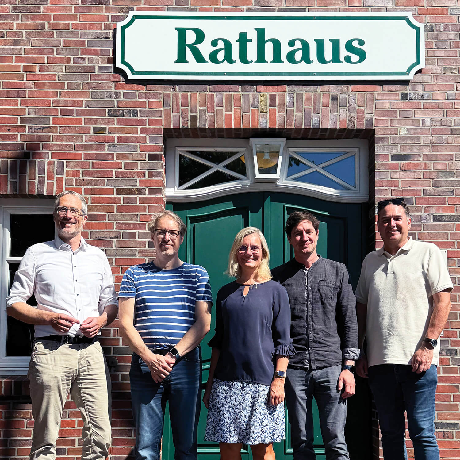 Fünf Erwachsene stehen nebeneinander vor einer Backsteinfassade mit grünem Doppeltor. Über der Tür hängt ein rechteckiges Schild mit grünem Rahmen und dem Wort 'Rathaus' in grüner Schrift auf weißem Grund. Die Personen tragen sommerliche Kleidung, darunter Hemden, ein gestreiftes T-Shirt und ein Kleid. Sonnenlicht wirft deutliche Schatten auf die Szene.