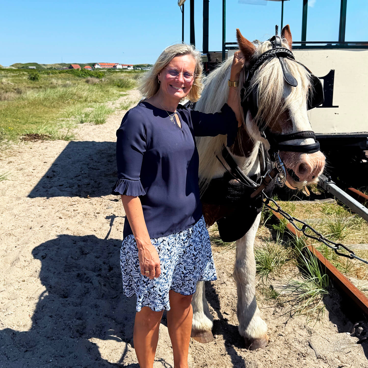 Eine Frau mit schulterlangem blondem Haar steht auf sandigem Boden neben einem weiß-braunen Pferd mit Zaumzeug. Sie trägt eine dunkelblaue Bluse mit halblangen Ärmeln und einen weißen Rock mit blauem Muster. Im Hintergrund ist ein helles, von Pferden gezogenes Fahrzeug mit großen Fenstern zu sehen. Die Szene spielt bei sonnigem Wetter im Freien, im Hintergrund sind Dünen und einige Gebäude erkennbar.