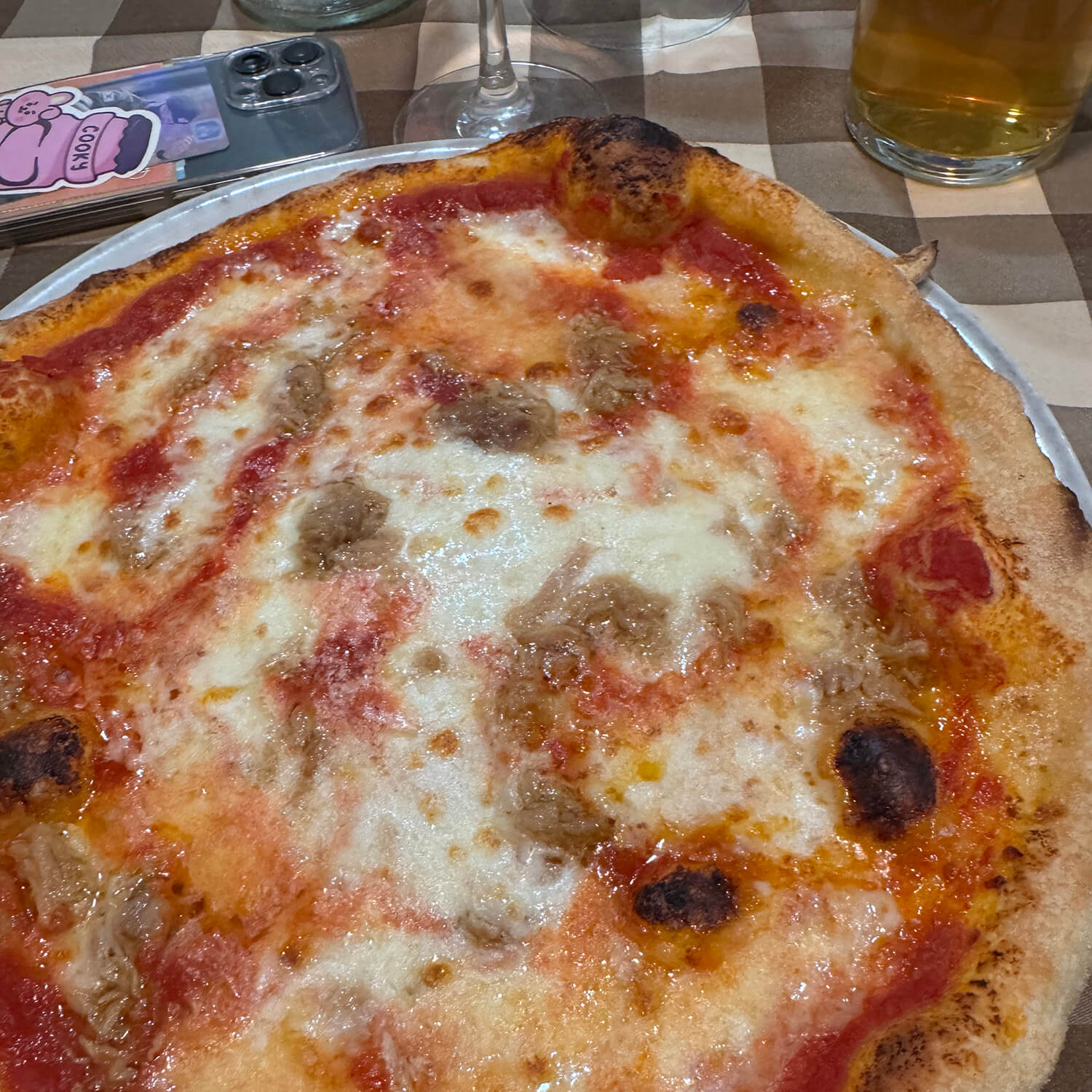Im Vordergrund liegt eine große Pizza mit dünnem, leicht gebräuntem Rand auf einem Teller. Die Pizza ist mit Tomatensauce, geschmolzenem Käse und einigen bräunlichen Belägen bedeckt. Im Hintergrund stehen ein Glas Bier, ein Weinglas und ein Smartphone mit einer rosa Hülle auf einem karierten Tisch.