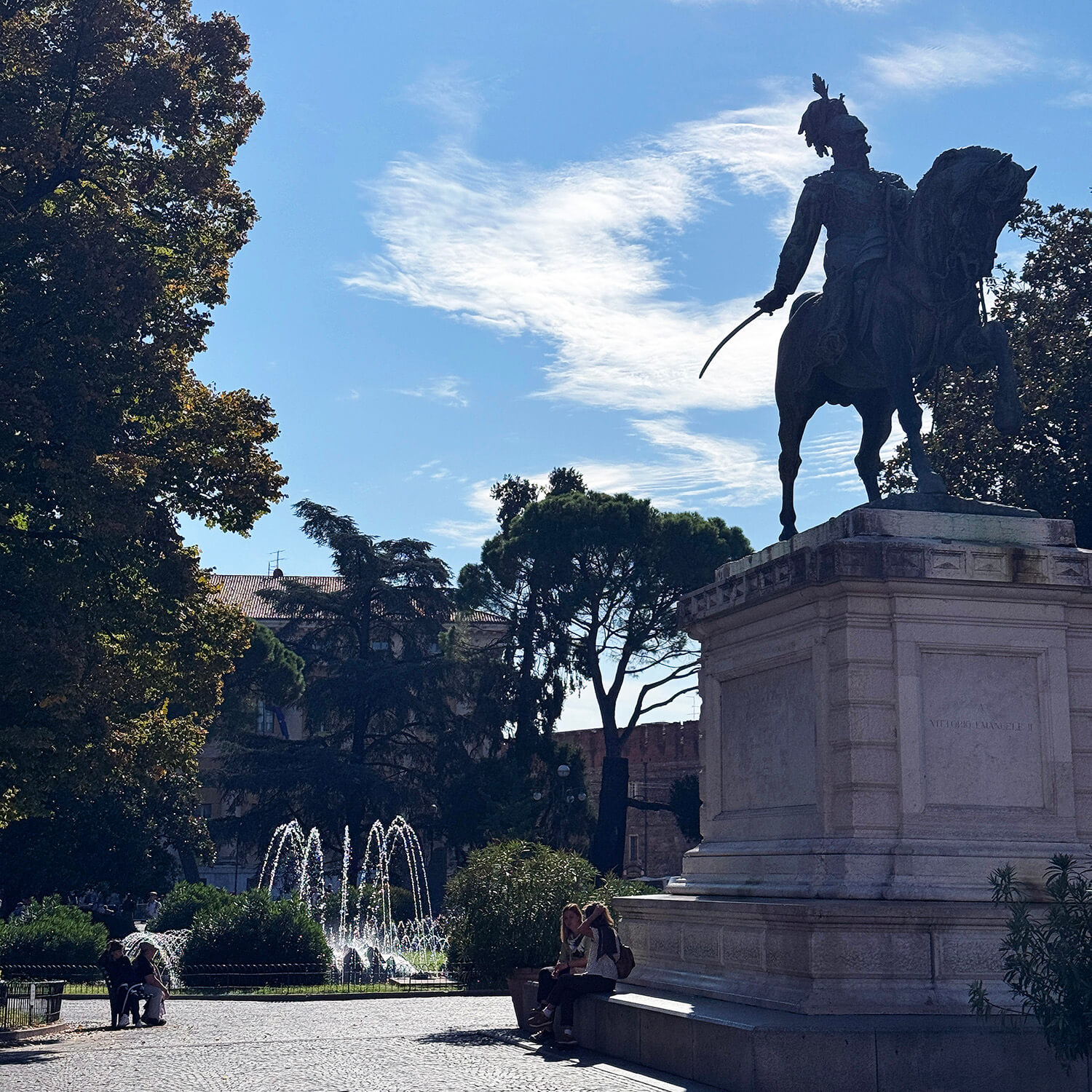 Im Vordergrund steht ein großes Reiterstandbild auf einem steinernen Sockel. Die Statue zeigt eine Figur mit erhobenem Schwert auf einem Pferd. Im Hintergrund sind mehrere Springbrunnen zu sehen, aus denen Wasserbögen aufsteigen. Hohe Bäume rahmen den Platz ein, und der Himmel ist leicht bewölkt. Auf einer Bank am Sockel der Statue sitzen zwei Personen.