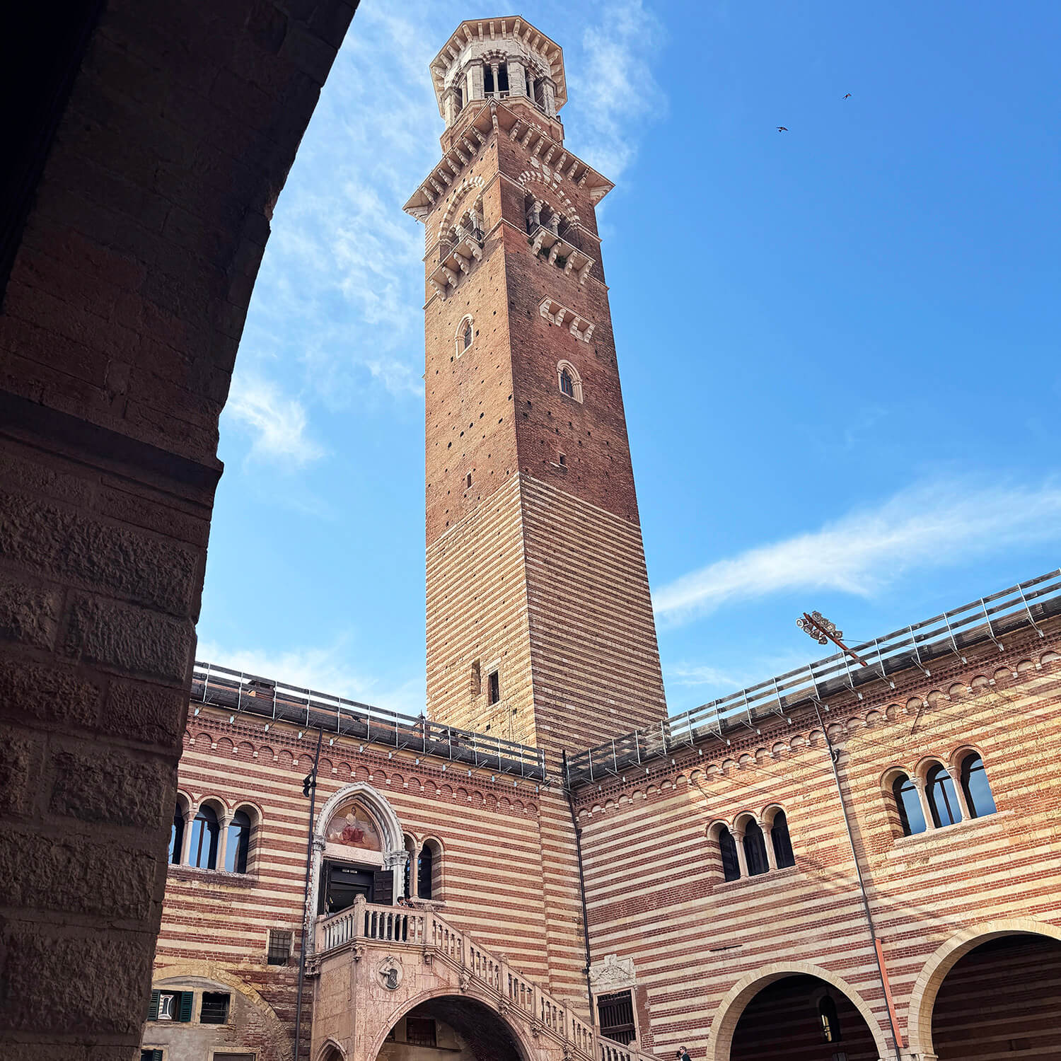 Im Mittelpunkt steht ein hoher, rechteckiger Turm aus rotbraunem Backstein mit hellen horizontalen Streifen. Der Turm besitzt mehrere kleine Fenster und eine dekorative Spitze mit Rundbogenöffnungen. Im Vordergrund ist ein Teil eines steinernen Bogens zu sehen, der den Blick auf den Innenhof mit Arkaden und einer breiten Treppe freigibt. Der Himmel ist klar und blau.