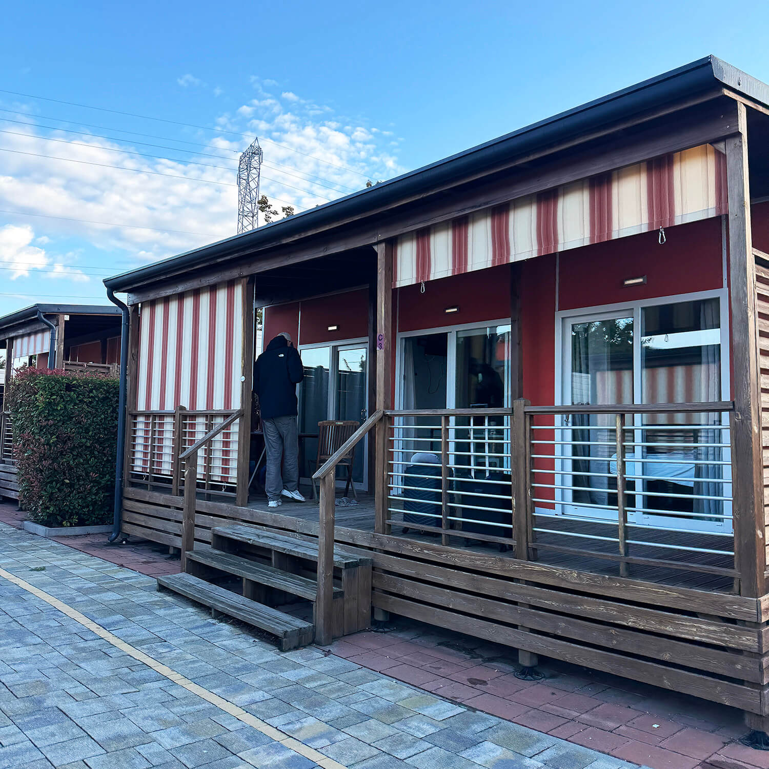Ein einstöckiges Gebäude mit roter Fassade und gestreifter Markise steht im Vordergrund. Die Terrasse ist mit einem Holzgeländer eingefasst und über eine kleine Treppe zugänglich. Zwei Personen stehen auf der Terrasse, eine davon an der Tür. Der Boden vor dem Gebäude ist mit hellen Pflastersteinen ausgelegt. Im Hintergrund ist ein weiterer, ähnlicher Gebäudeteil zu sehen. Der Himmel ist blau mit einigen Wolken.