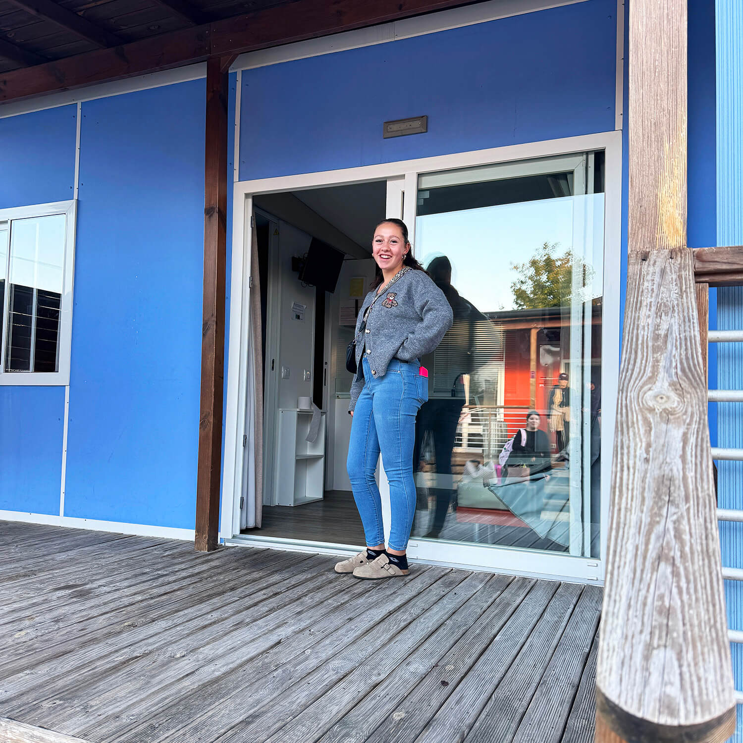 Eine junge Frau steht auf einer Holzterrasse vor einer blauen Gebäudewand mit großen Fenstern und einer offenen Glastür. Sie trägt eine graue Jacke, blaue Jeans und helle Schuhe. Im Hintergrund sind ein Badezimmer und Spiegelungen im Glas sichtbar. Die Szene wirkt freundlich und entspannt.