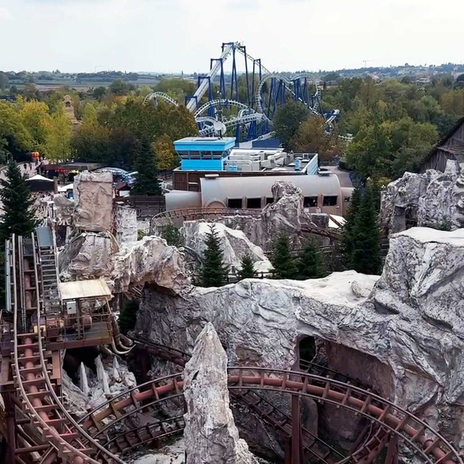 Achterbahnschienen schlängeln sich durch eine felsige Landschaft mit künstlichen Felsen und Tannen. Im Hintergrund sind weitere Fahrgeschäfte und eine große, blau-weiße Achterbahn zu sehen. Dichte Bäume umgeben das Gelände, dahinter erstreckt sich eine flache Landschaft.